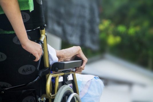 kompleksowa pomoc, osoba starsza na wózku inwalidzkim pchana przez opiekuna